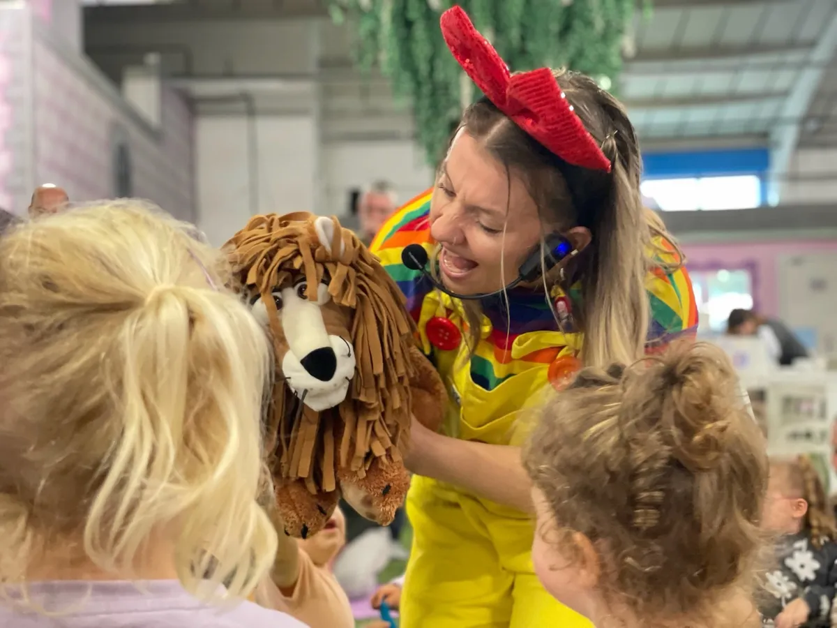 Coralie in her rainbow outfit and red bow, roaring along with a lion puppet for a group of toddlers